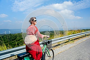 Back of woman with sunglasses stand after ride the bicycle on road on the mountain near row of wind turbines or windmill and she