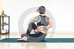 Back woman with tattoos in ardha matsyendrasana yoga pose in front of a white wall with yoga elements