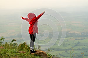 Back of woman wear red coat and cover with hood and stand with spread her arms near cliff on the mountain and look forward to