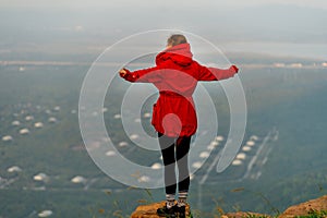 Back of woman wear red coat and stand with spread her arms near cliff on the mountain and look forward to beautiful view with warm