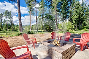 Back yard with fire pit and red chairs near newly bild luxury real estate home with forest biew and green grass