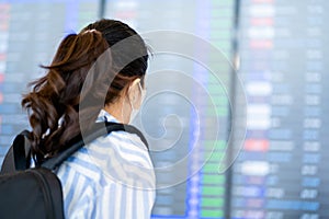Back of young asian woman traveler with backpack wearing a protective mask against the virus and looking at departure board in