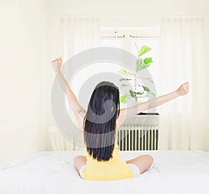 Back of young asian woman wearing yellow undershirt exercising yoga while sitting on a white bed by the window with a thin curtain