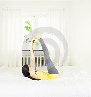 Back of young asian woman wearing yellow undershirt exercising yoga while sitting on a white bed by the window with a thin curtain