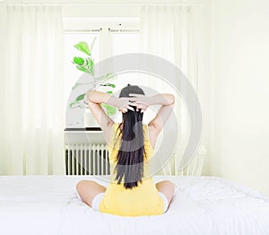 Back of young asian woman wearing yellow undershirt exercising yoga while sitting on a white bed by the window with a thin curtain