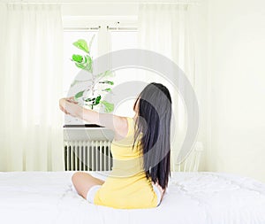 Back of young asian woman wearing yellow undershirt exercising yoga while sitting on a white bed by the window with a thin curtain