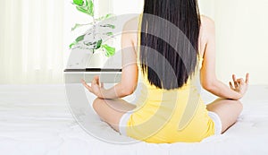 Back of young asian woman wearing yellow undershirt exercising yoga while sitting on a white bed by the window with a thin curtain