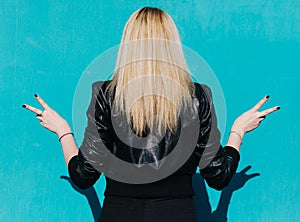 Back of a young beautiful blonde girl on the background of turquoise wall sunny summer day shows victory sign, outdoor, filter.