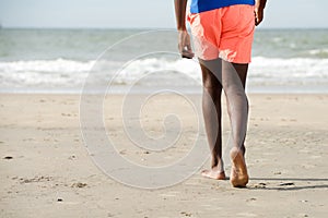 Back of young black man walking barefoot on beach towards water