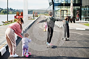 Back of young stylish mother with four kids outdoor. Sports family spend free time outdoors with scooters and skates