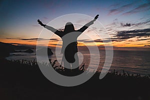 Back of a young traveler girl standing on on the edge of a cliff by the ocean coast watching a beautiful sunset. Arms wide open.