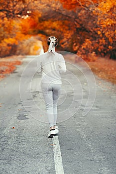 Back of young woman in sportswear jogging on asphalt road on background of red foliage, girl engaged in sport outdoors on autumn