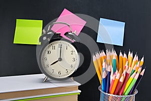 On the background of a black Board with stickers, there is an alarm clock on the books and a glass with colored pencils