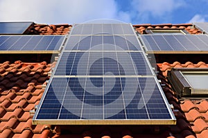 Background and close-up of a solar panel on a roof of a family home with red roof tiles, against a blue sky with clouds and