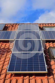 Background and close-up of a solar panel on a roof of a family home with red roof tiles, against a blue sky with clouds and