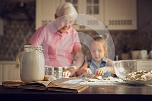 Background image cookbook, table, rolling pin, focus on the foreground. Girl and her grandmother cooking on kitchen.