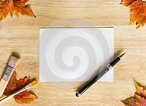 Background texture with wooden table and autumnal leaves. Frame, made from pen, painting brushes, autumn leaves and white paper