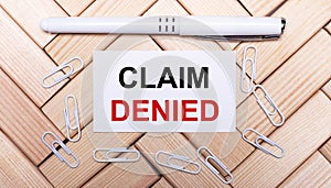 On a background of wooden blocks, a white pen, white paper clips and a white card with the text CLAIM DENIED. View from above