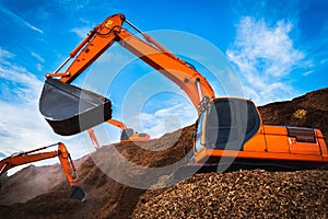 Backhoe standing on big pile of wood chips loading up into truck. Woodchips raw material storing and transportation.