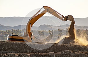 Backlit Backhoe scooping dirt and dust with hills in the background