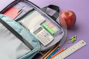 Backpack with fresh apple, calculator and stationery on lilac background