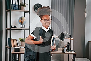 With backpack and notepad. Cute little girl in school uniform is standing indoors in domestic room