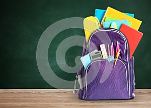 Backpack with school stationery on wooden table near green chalkboard, space for text
