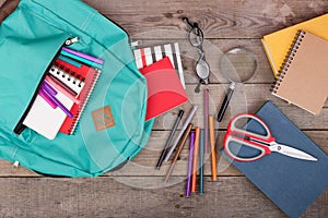 Backpack and school supplies: books, magnifying glass, notepad, felt-tip pens, eyeglasses, scissors on wooden table