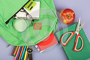 Backpack and school supplies: magnifying glass, notepad, felt-tip pens, eyeglasses, scissors, calculator, book, watch on blue pape