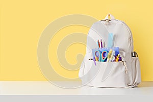 Backpack with school supplies on white table near yellow wall