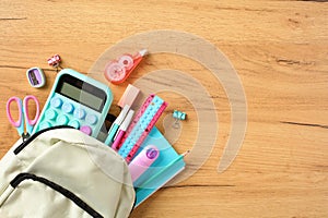 Backpack with school supplies on wooden table. Calculator, ruler, notepad, sharpener, proofreader, pens inside bag