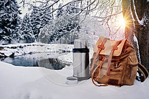 Backpack of traveller, thermos and enameled mug of coffee or tea against snowy winter landscape background
