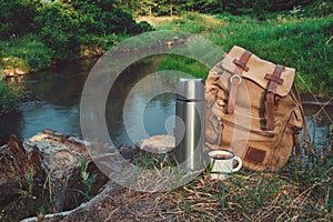 Backpack of traveller, thermos and Enameled mug of coffee or tea on bank of beautiful forest river