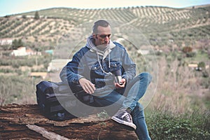 Backpacked adventurer traveler man, sitting on log in forest against mountains, drinking coffee with relaxing on nature