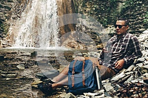 Backpacker Man Drink Tea Or Coffee From Thermos On Waterfall Background Resting Camping Destination Travel Concept