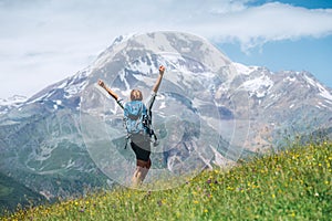 Backpacker woman rising arms and greeting snowy slopes of Kazbek 5054m mountain with a backpack while she walking by green grass