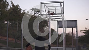 Backside footage of a young basketball female player jogging with ball on the outdoors court and throw it to the hoop