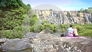 Backside girl hugs guy on stone against waterfall