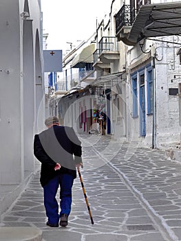 Backside of unrecognizable elderly senoir man wearing beret hat holding a cane walking in the small mediterranean alley of Paros