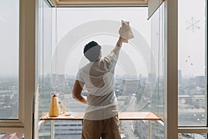 Backside view of asian Thai man wiping window glass in room apartment with city view, keep glass sparking clean