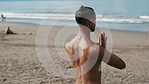 Backside view athletic shirtless man doing yoga on beach near ocean