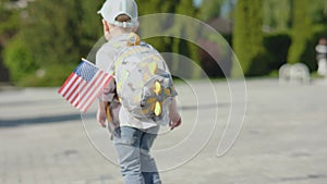 backside view baby boy in blue cap, grey backpack with waving US flag walking