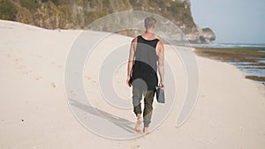 Backside view, lonely man with yoga mat in hands walks along sandy beach