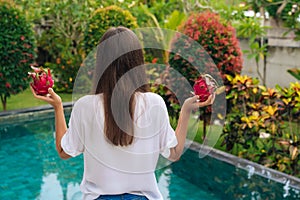 Backside view of slender girl standing with dragon fruits, pitaya near pool in beautiful garden