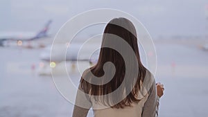 Backside view of woman standing in terminal of airport, looks at plane out window