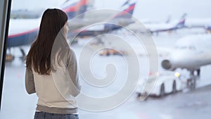 Backside view of woman standing in terminal of airport, looks at plane out window