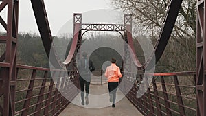 Backside view of young sportive couple running across bridge in parkland. Woman and man athletes in bright sportswear jogging outd