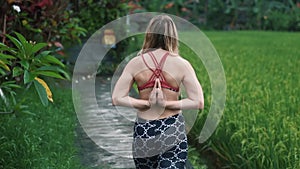 Backside view young woman doing yoga exercises outdoors, greenery on background
