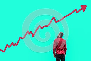 Backside of young bearded handsome businessman in red shirt standing and looking at growing graph chart analysis diagram drawed on