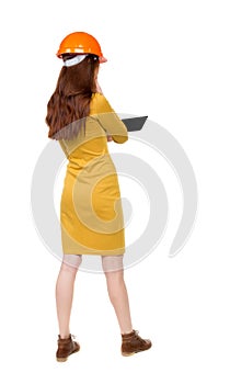 Backview of business woman in construction helmet stands and enjoys tablet or using a mobile phone.